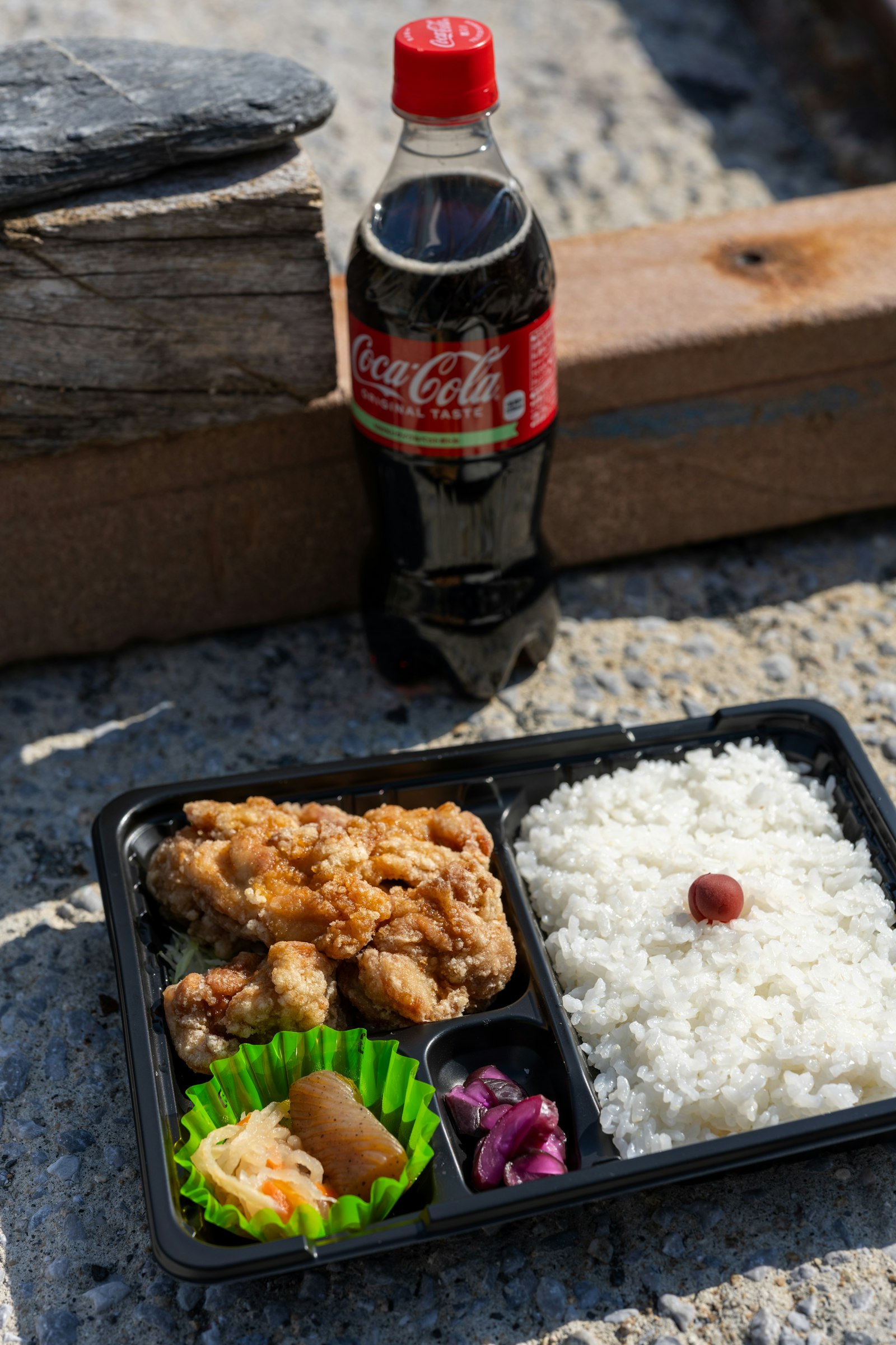 A karaage chicken bento with rice, pickles, and a Coca-Cola — set on weathered stone like takeaway from a North Shore plate lunch joint.